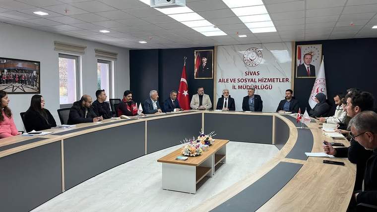 Toplantı salonunda uzun masanın çevresinde oturan kişiler, duvarda Türk bayrağı ve kurum afişiyle görülüyor.