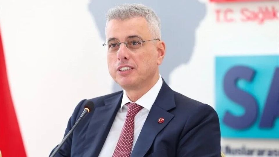 Mikrofon önünde konuşan gözlüklü bir kişi, arka planda “T.C. Sağlık” yazılı pano bulunuyor.