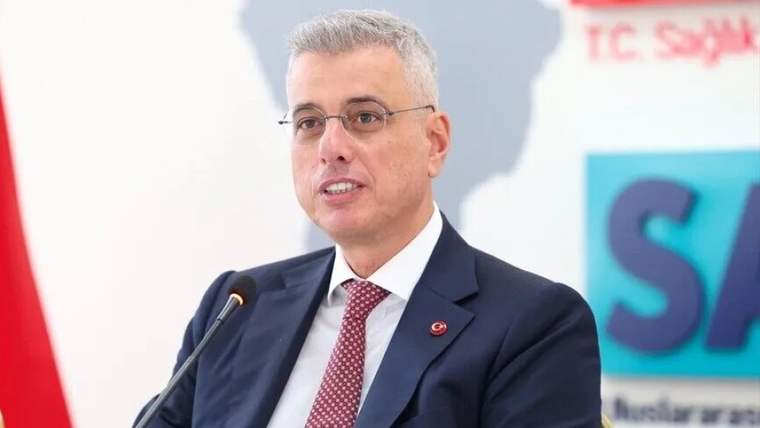 Mikrofon önünde konuşan gözlüklü bir kişi, arka planda “T.C. Sağlık” yazılı pano bulunuyor.