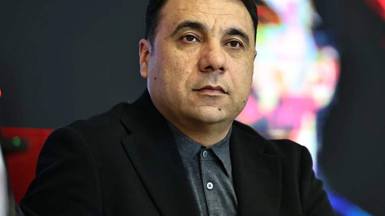 Koyu renk ceket ve gri gömlek giyen bir kişi, renkli ışıkların bulunduğu kapalı bir ortamda oturuyor.