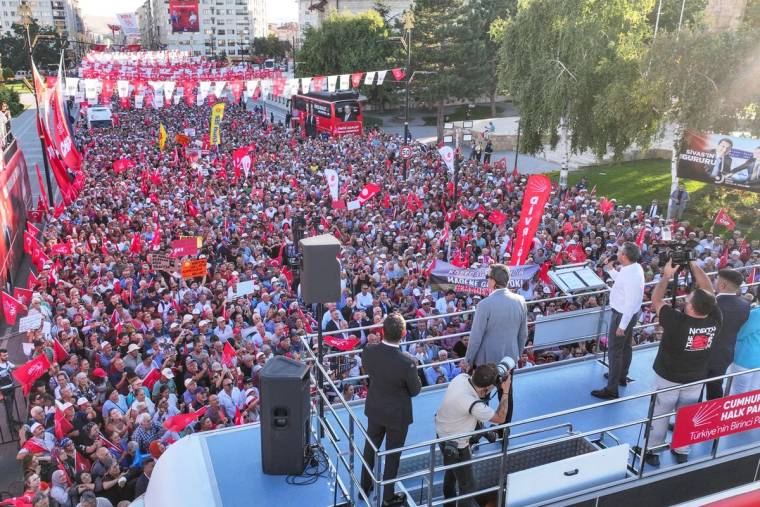 Bir platformda konuşan kişiler, kalabalık bir topluluğa hitap ediyor. Arka planda bayraklar ve afişler var.