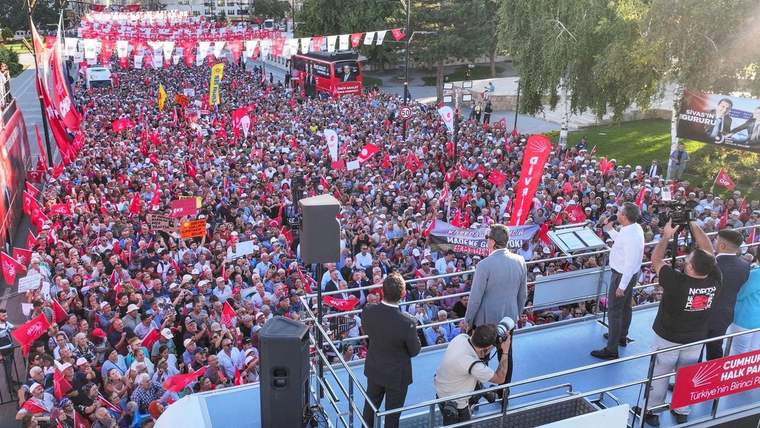 Bir platformda konuşan kişiler, kalabalık bir topluluğa hitap ediyor. Arka planda bayraklar ve afişler var.