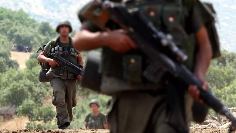 Askeri üniforma giymiş birkaç asker, yürüyerek ilerliyor. Arkada yeşil ağaçlar ve açık bir alan görünmekte.