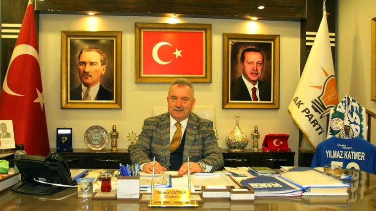 Bir masa başında oturan bir kişi, arkasında Türk bayrağı ve iki portre var.