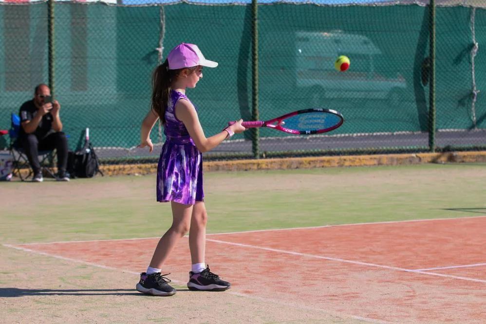 Tenis kortunda, mor elbiseli bir çocuk tenis raketiyle topa vurmak üzere duruyor.