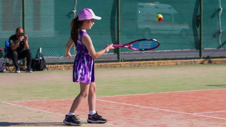 Tenis kortunda, mor elbiseli bir çocuk tenis raketiyle topa vurmak üzere duruyor.