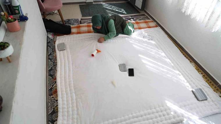 Odanın zemininde serili beyaz yorganın üzerinde yeşil kıyafetli bir kişi, yanında telefonlarla görülüyor.