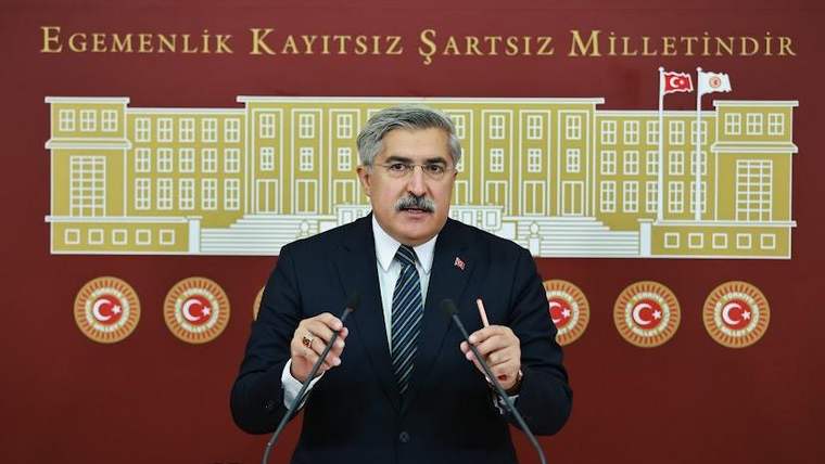 Kravatlı bir kişi, Türkiye bayrakları ve meclis çiziminin önünde kürsüde konuşuyor.
