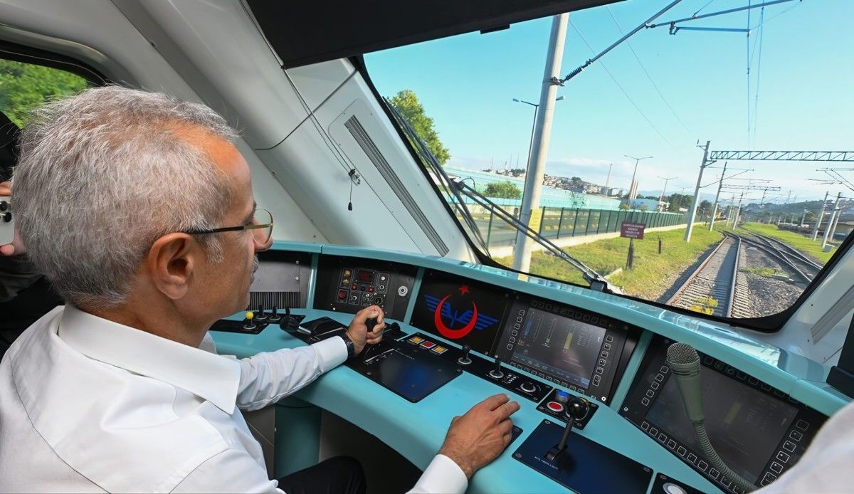 Tren sürücü kabininde oturan bir kişi, kontrol ekranları ve kumanda paneli önünde rayları izliyor.