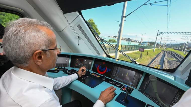 Tren sürücü kabininde oturan bir kişi, kontrol ekranları ve kumanda paneli önünde rayları izliyor.