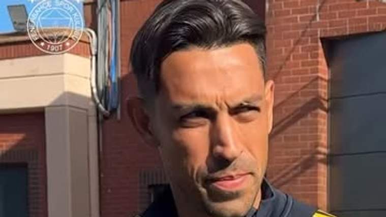 Sarı lacivert spor kıyafetli bir kişi, elinde kâğıt bardakla tuğla bina önünde duruyor.