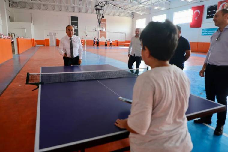 Kapalı spor salonunda bir çocuk masa tenisi masasının başında dururken çevresinde yetişkinler görülüyor.