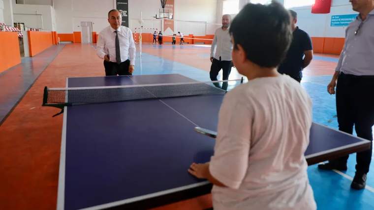 Kapalı spor salonunda bir çocuk masa tenisi masasının başında dururken çevresinde yetişkinler görülüyor.
