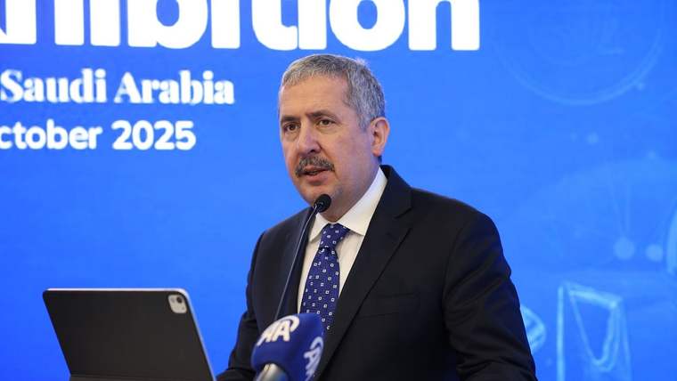 Mikrofonların önünde kürsüde konuşan takım elbiseli bir kişi, mavi fonda tabletle birlikte görülüyor.