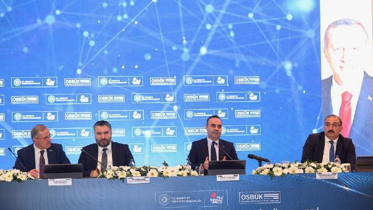 Masada oturan dört erkek ve arkalarında toplantı logoları bulunan panoda “Anadolu Organize Sanayi Bölgeleri İstişare Toplantısı” yazısı görülüyor.
