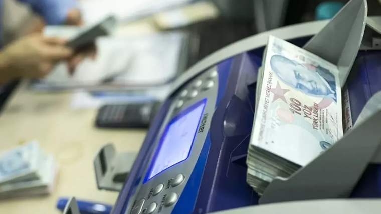Banknot sayma makinesinin haznesinde Türk lirası banknotları, arka planda masa başında çalışan biri görülüyor.