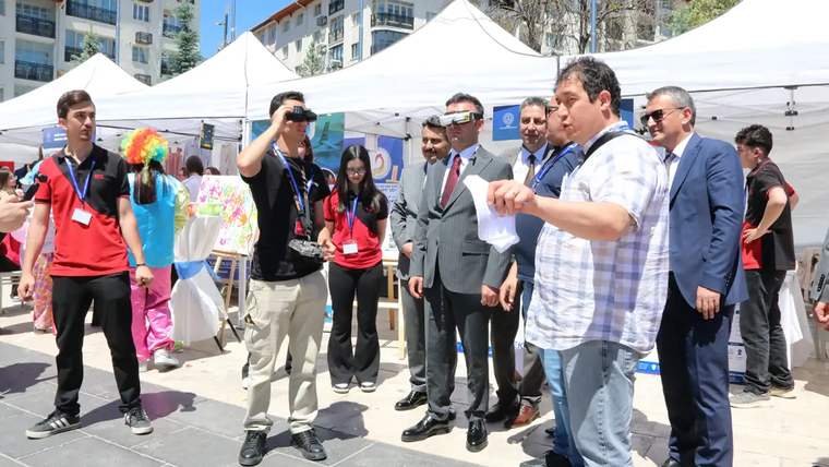 Açık hava stantları önünde bir kişi elindeki kâğıdı tutup el hareketi yaparken çevresinde insanlar duruyor.