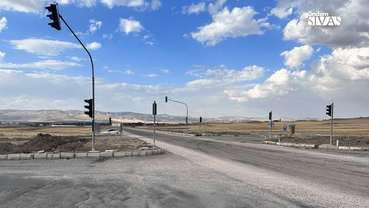 Trafik ışıkları bulunan geniş kavşakta asfalt ve toprak yollar, çevredeki tepelerle birlikte görülüyor.