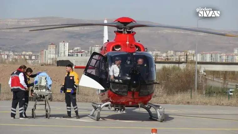 Kırmızı bir helikopter pistte dururken iki görevli yanında tekerlekli sedyeyi taşıyor; kabinde iki kişi görülüyor.
