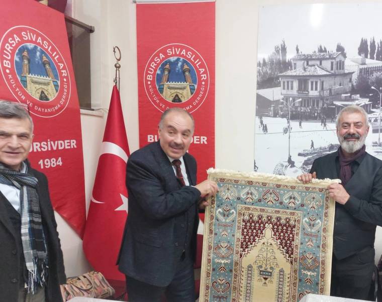 Bir grup erkek, elinde bir halı tutarak gülümsüyor; arka planda Türk bayrağı ve Sivas ile ilgili afişler var.