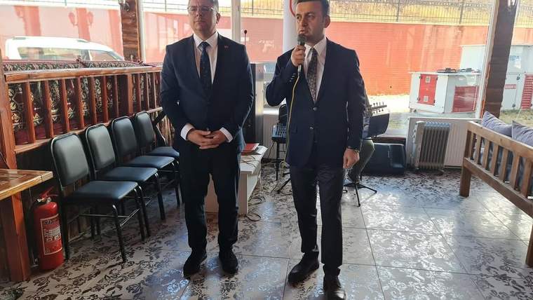 Kahvaltı programında konuşma yapan iki kişi, takım elbise giymiş.