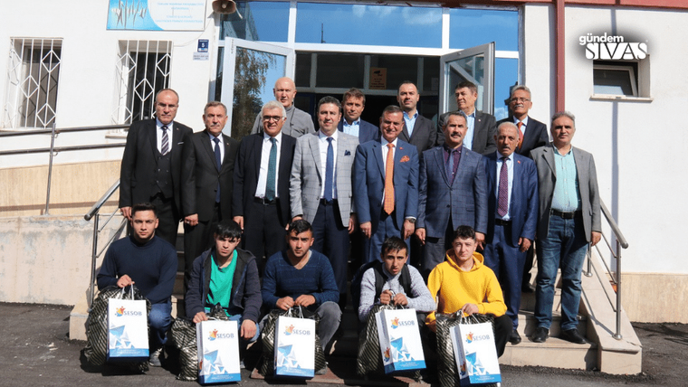 Bir binanın girişinde takım elbiseli erkekler ve gençler birlikte duruyor; öndekiler logolu poşet tutuyor.