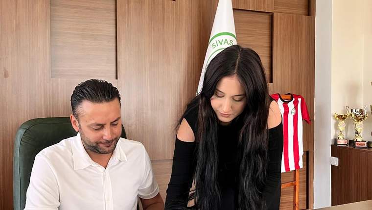 Bir kadın, masada oturan bir erkeğin yanında kırmızı dosyadaki belgeyi imzalıyor; arkada kupa ve formalar bulunuyor.