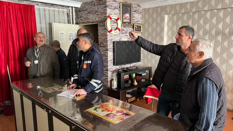 Bir odada beş erkek, masa çevresinde duruyor; siyah montlu biri duvardaki noktayı gösteriyor.