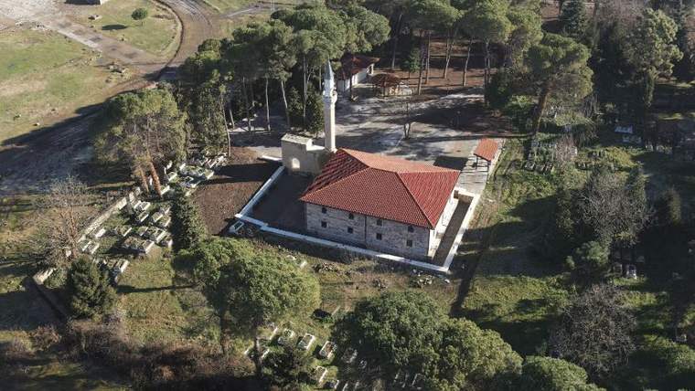Ağaçlar ve mezar taşlarıyla çevrili, kırmızı çatılı taş cami ile minaresi havadan görülüyor.