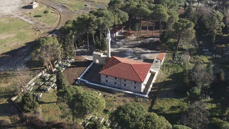 Ağaçlık alanda, yanında mezarlar bulunan kırmızı çatılı minareli bir cami binası havadan görülüyor.