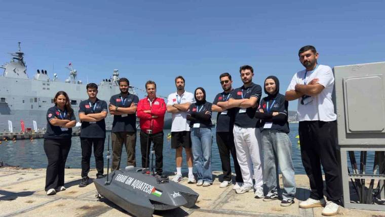 Liman kenarında bir grup insan, üzerindeki yazılar görülen küçük teknenin yanında duruyor; arkada büyük bir gemi var.