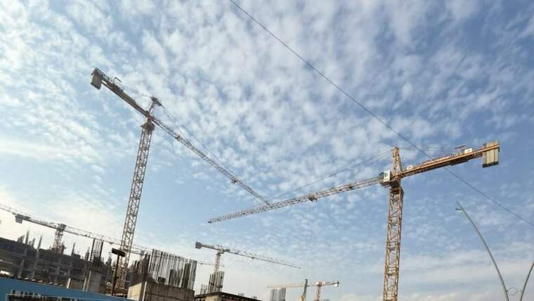 Gökyüzü altında, betonarme bir yapının çevresinde yükselen üç kule vinç ve inşaat demirleri görülüyor.