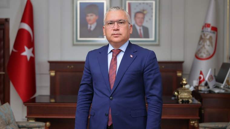 Takım elbiseli bir kişi, Türk bayrağı ve duvarındaki portrelerin bulunduğu makam odasında duruyor.