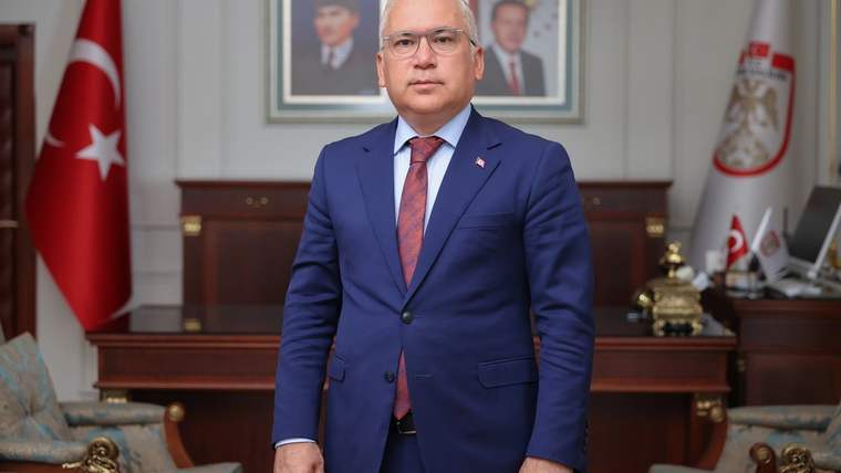 Takım elbiseli bir kişi, Türk bayrağı ve duvarındaki portrelerin bulunduğu makam odasında duruyor.