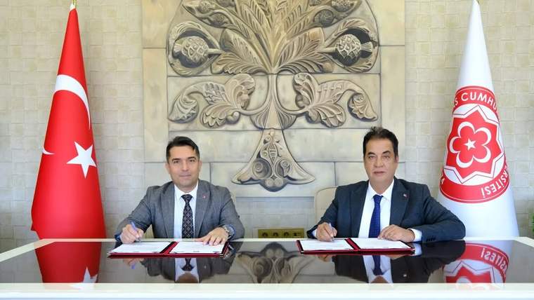 Türk bayrağı ve üniversite logolu bayrak önünde iki kişi masada belgeleri imzalıyor.