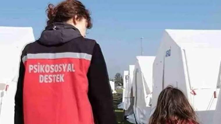 Sırtında “Psikososyal Destek” yazılı kırmızı yelekli kişi, beyaz çadırların arasında bir çocukla yürüyor.