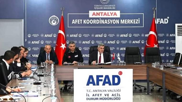 Toplantı salonunda Türk bayrakları önünde masada oturan dört kişi ve çevresindeki katılımcılar görülüyor.