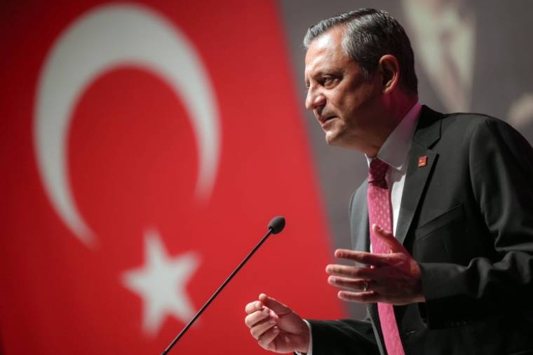 Mikrofon önünde konuşan takım elbiseli bir kişi, arka planda Türk bayrağıyla görülüyor.