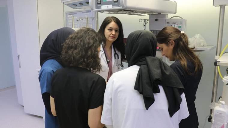 Beyaz önlük giymiş bir doktor, etrafında dört kişiyle birlikte bir inkübatörün önünde duruyor.