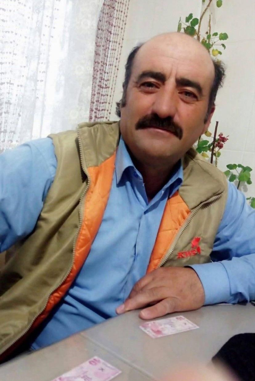 Bir adam, mavi gömlek ve krem rengi yelek giymiş, gülümseyerek kameraya bakıyor.