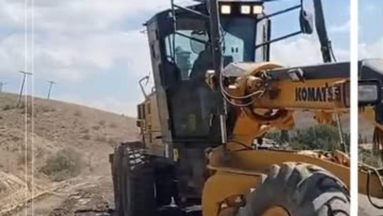 KOMATSU marka greyder, toprak yolda çalışırken yanında kurak ve engebeli arazi görülüyor.