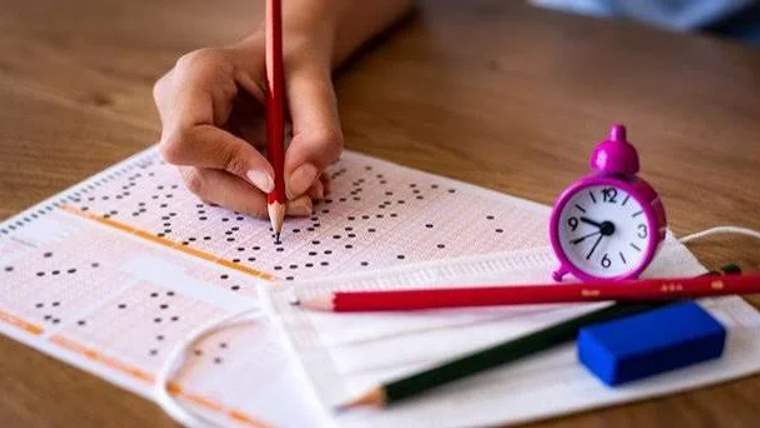 Bir el, üzerinde siyah noktalar bulunan bir kağıda kırmızı kalemle işaretler yapıyor; yanında masa saati ve kalemler var.