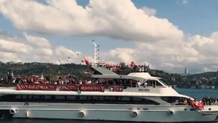 Kırmızı ve sarı bayraklar taşıyan kalabalığın bulunduğu yolcu teknesi, denizde kıyı açıklarında görülüyor.