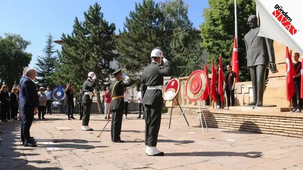 Askerler, Atatürk heykelinin önünde selam duruyor; arka planda Türk bayrakları ve çiçekler var.