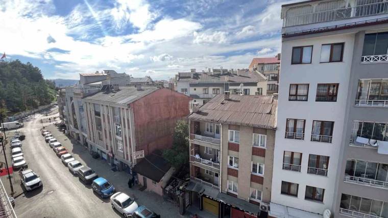Apartman binaları arasında alçak çatılı bir yapı, caddede park etmiş araçlar ve uzakta ağaçlık alan görülüyor.