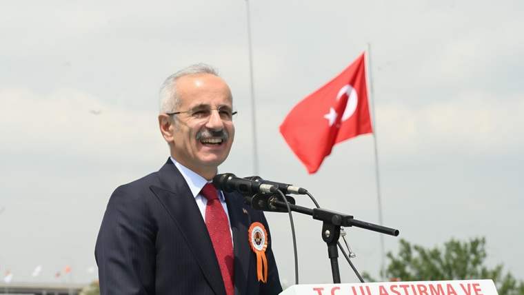 Mikrofon önünde konuşan bir kişi, üzerinde rozet var ve arka planda Türk bayrağı dalgalanıyor.