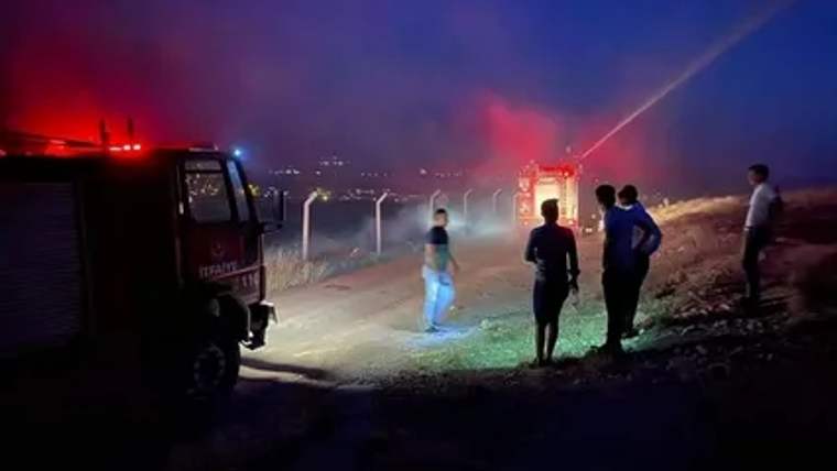 Gece karanlığında kırmızı ışıkları yanan bir itfaiye aracı, dumanlı alanda duran insanlara bakıyor.