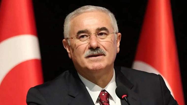 Kürsüde konuşan bir kişi, arkasında Türk bayrağı bulunan bir sahnede yer alıyor.