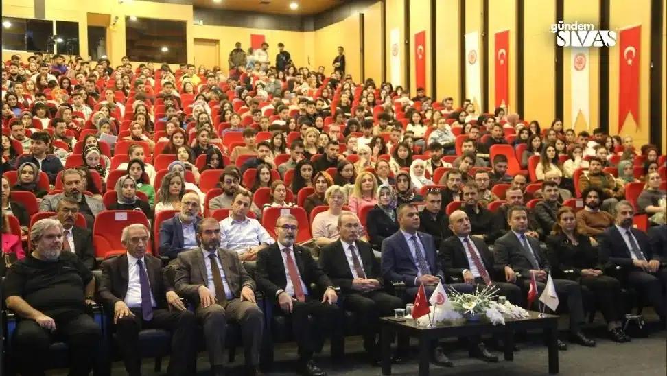 Bir konferans salonunda, dinleyicilerle birlikte sahnede oturan konuşmacılar yer alıyor.