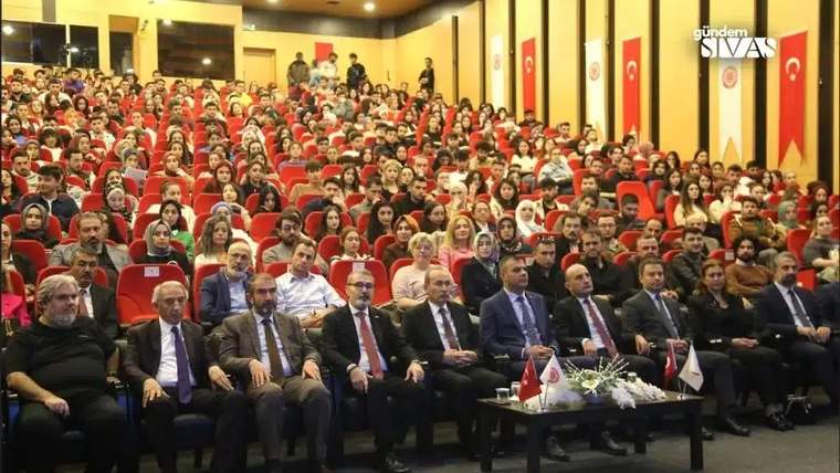 Bir konferans salonunda, dinleyicilerle birlikte sahnede oturan konuşmacılar yer alıyor.
